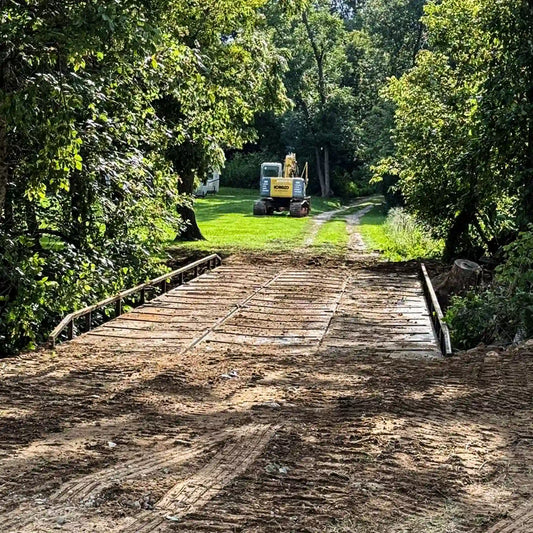 Yellow excavator crossing a modular steel construction bridge over a creek, showing the bridge carrying heavy equipment loads.