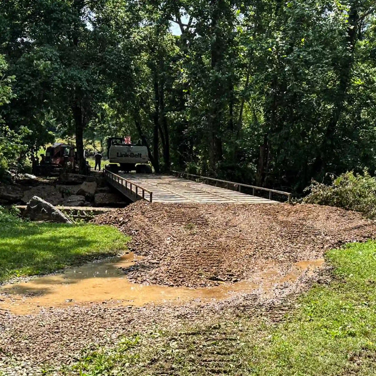 Front view of a modular steel construction bridge set into a dirt access road, with an excavator visible beyond the crossing.