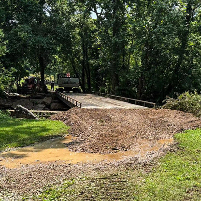 Front view of a modular steel construction bridge set into a dirt access road, with an excavator visible beyond the crossing.