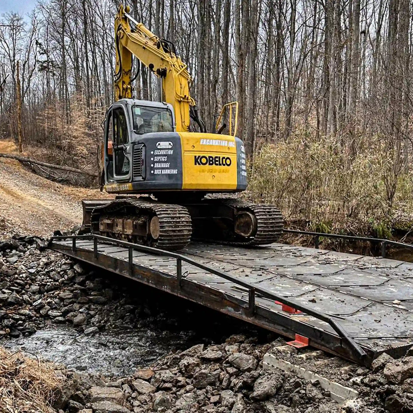 Compact excavator positioned on a steel construction bridge over a creek, demonstrating equipment access on a jobsite.