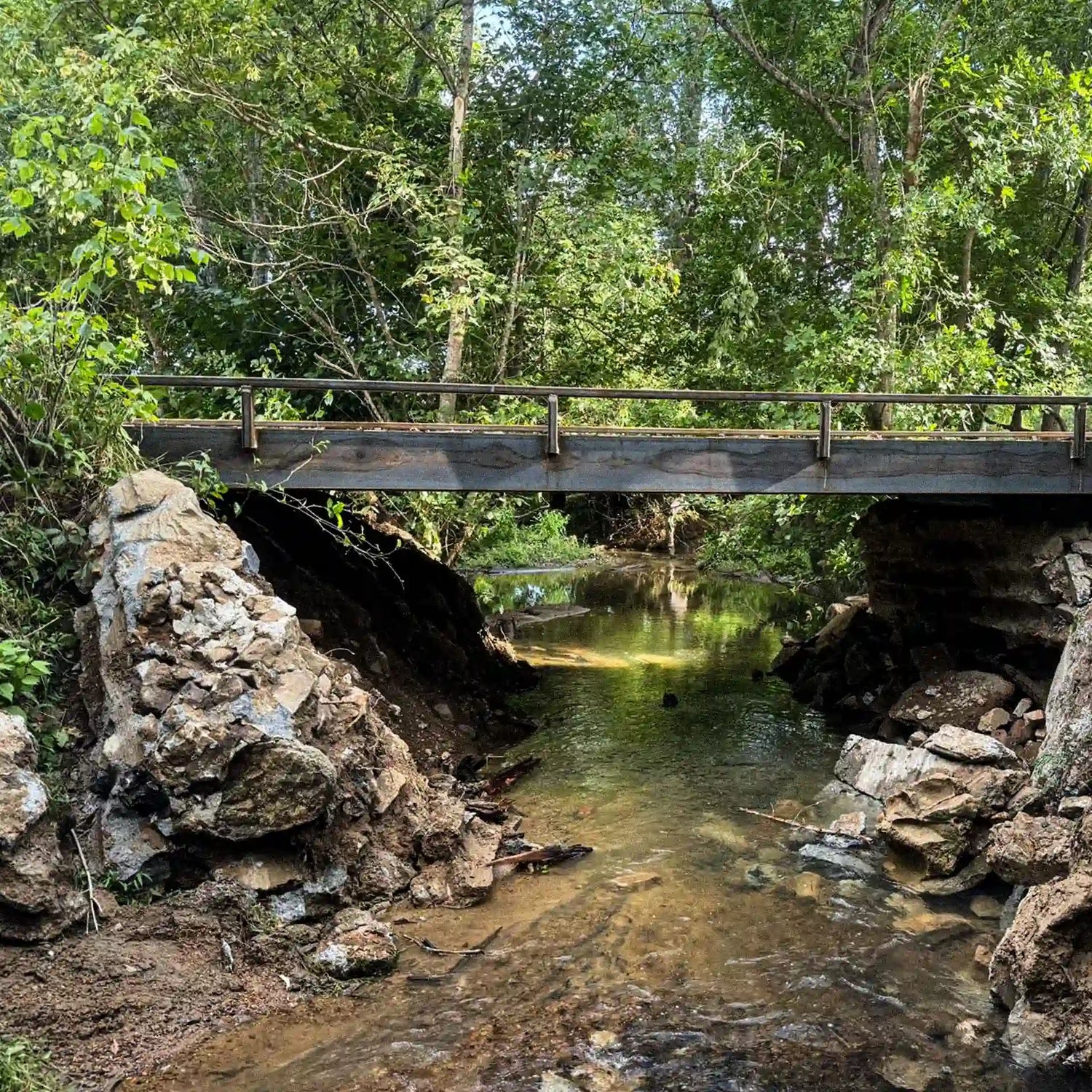 Steel logging bridge installed over a shallow creek with stone abutments and side railings in a wooded area.