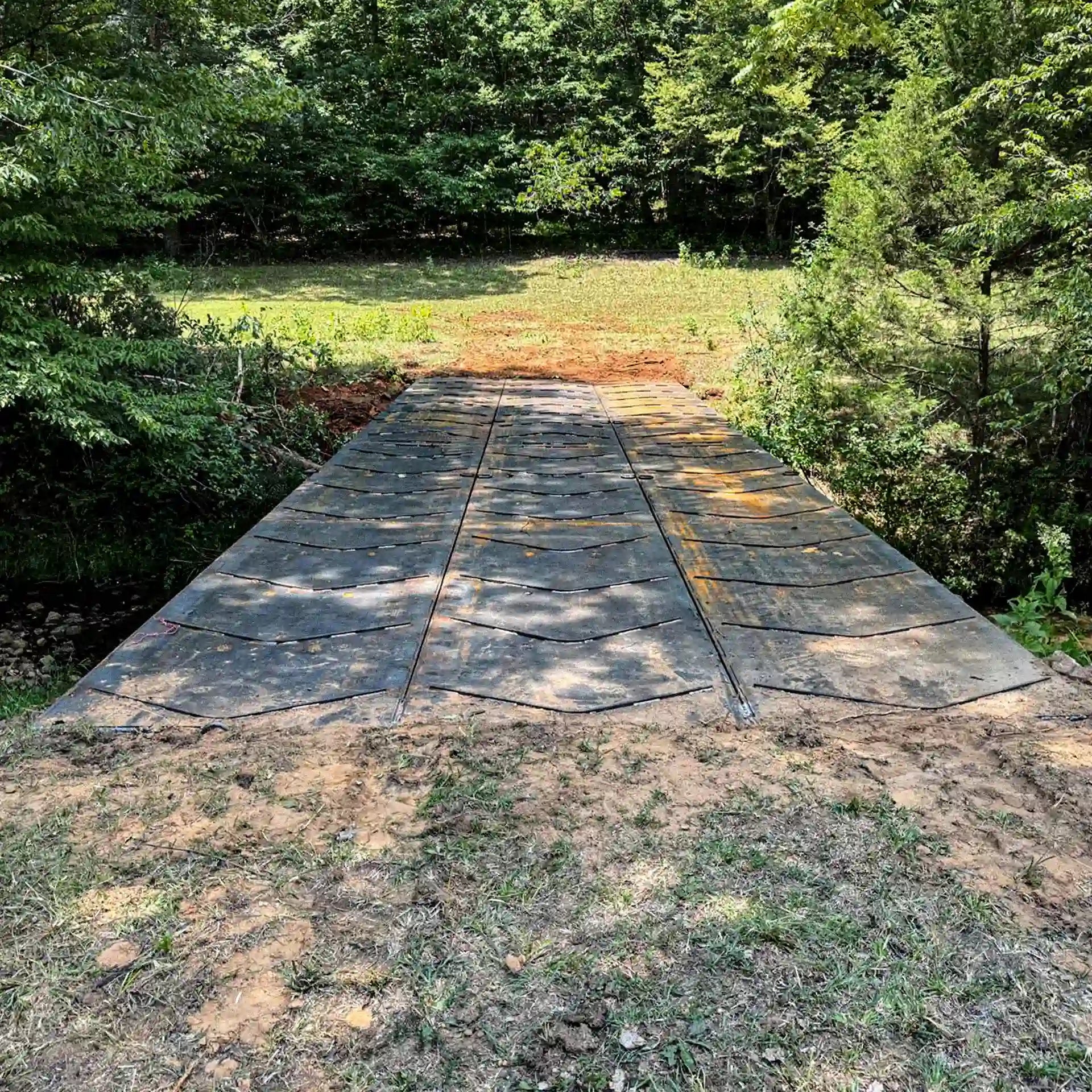 Front view of a modular steel logging bridge spanning a small creek, shown installed between dirt approaches in a wooded clearing.