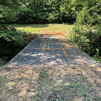 Front view of a modular steel logging bridge spanning a small creek, shown installed between dirt approaches in a wooded clearing.