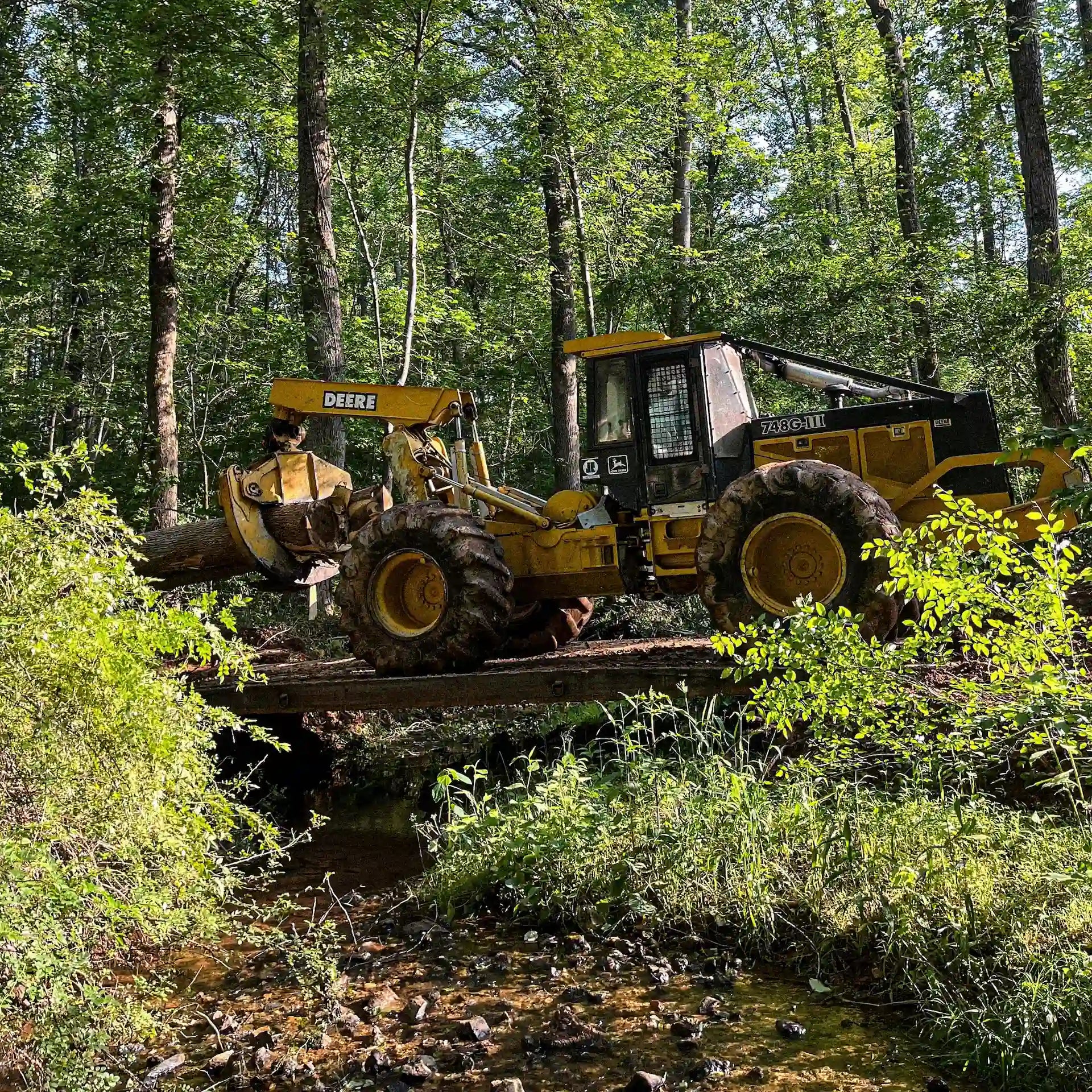 Side view of a John Deere skidder carrying a log across a modular steel logging bridge over a small stream.