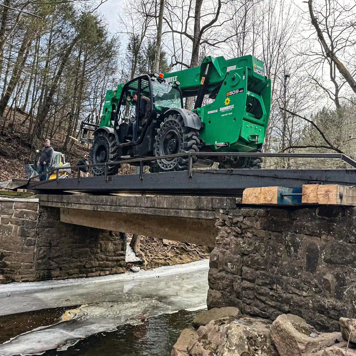Green telehandler driving across a steel logging bridge over a creek, demonstrating heavy equipment load capacity.
