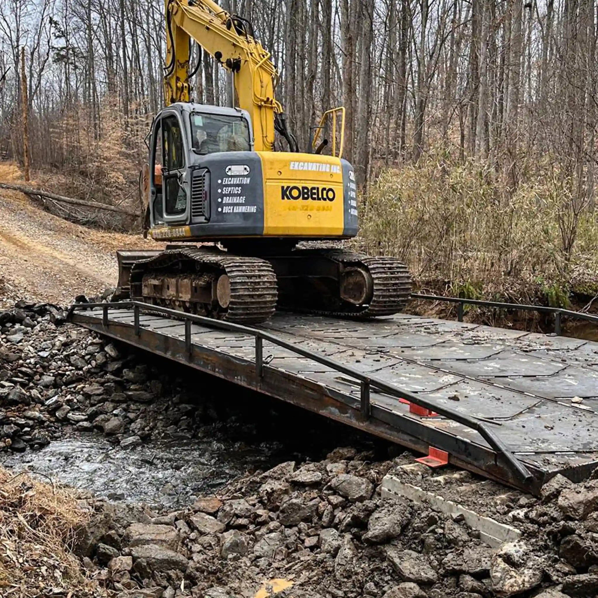 Kobelco excavator on a portable steel bridge over a stream in a forested area