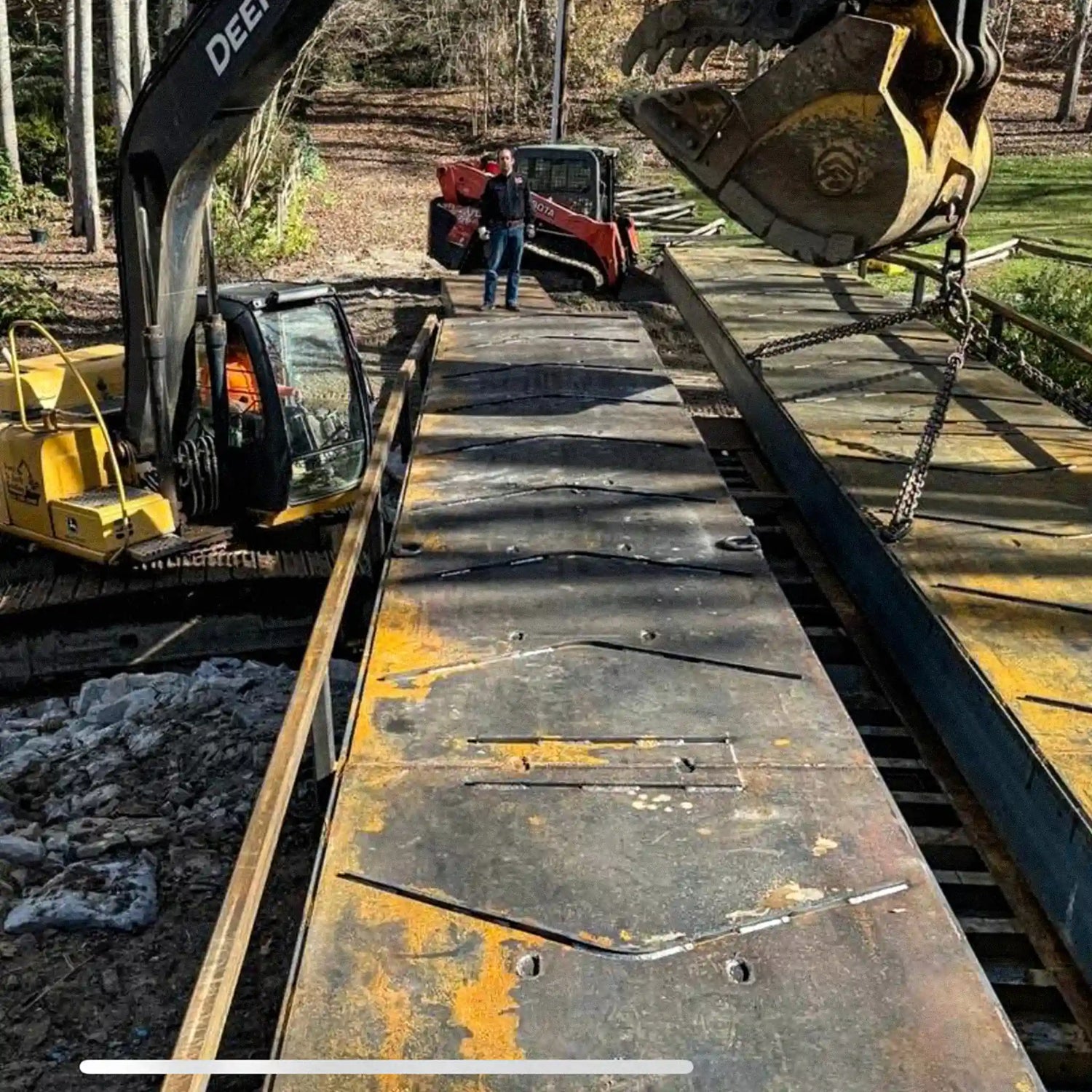 excavator installing a component of a portable steel bridge with a man standing in the background