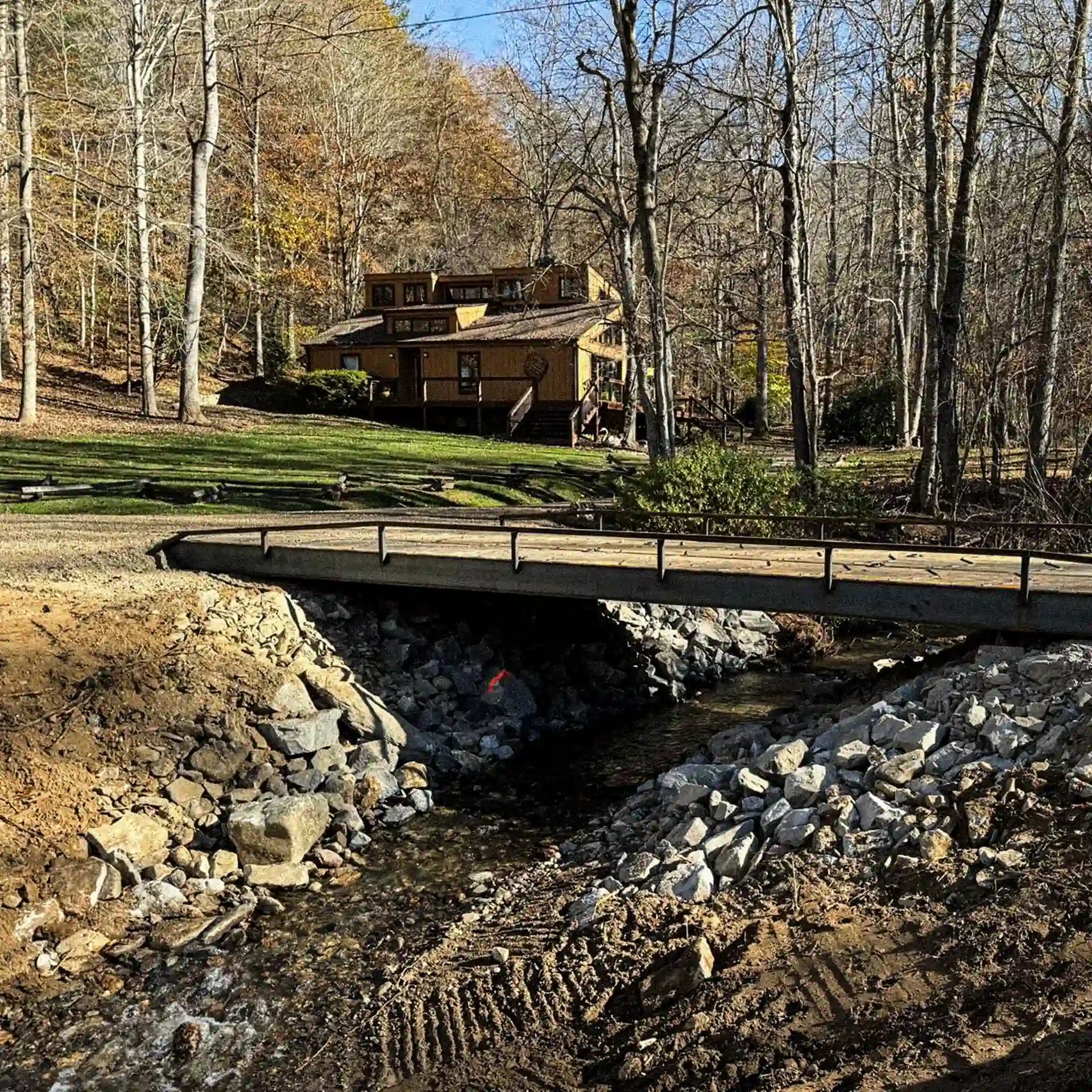 Steel residential bridge over a small stream on a wooded property, providing driveway access to a house in the background.