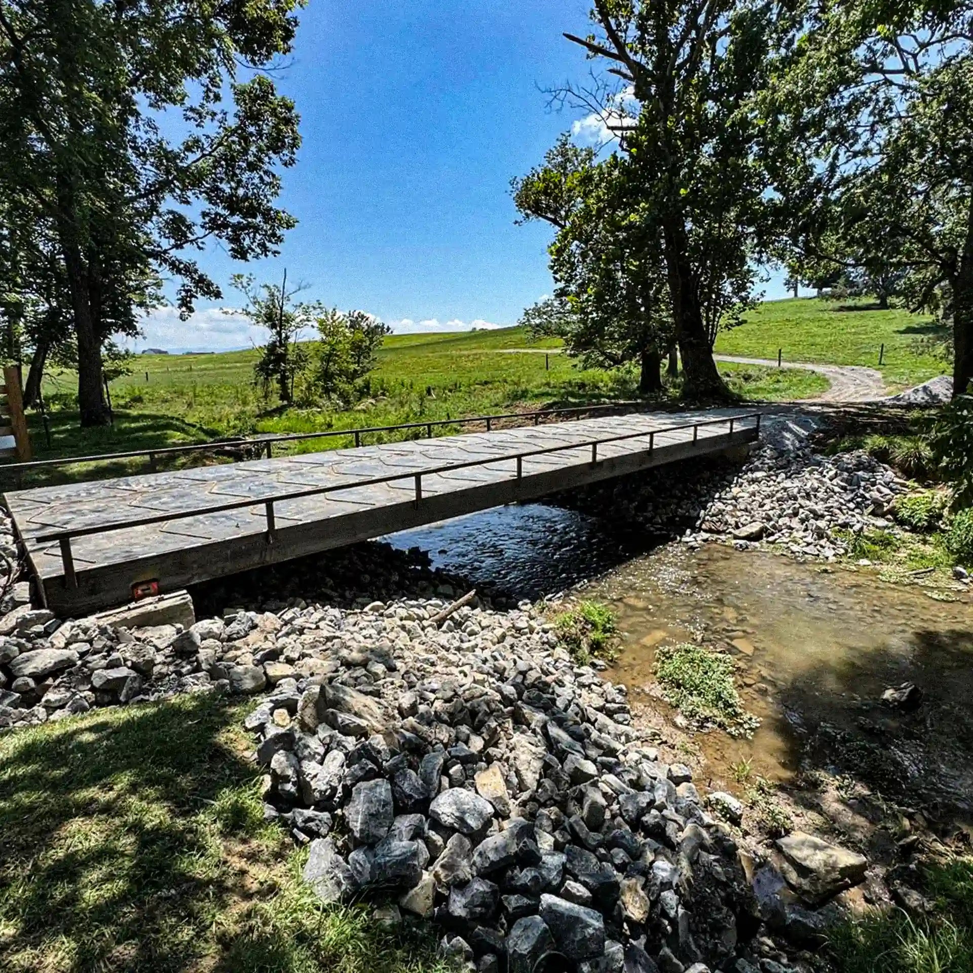 Angled side view of a residential steel bridge crossing a small creek in open pasture, with rock approaches and mature trees.