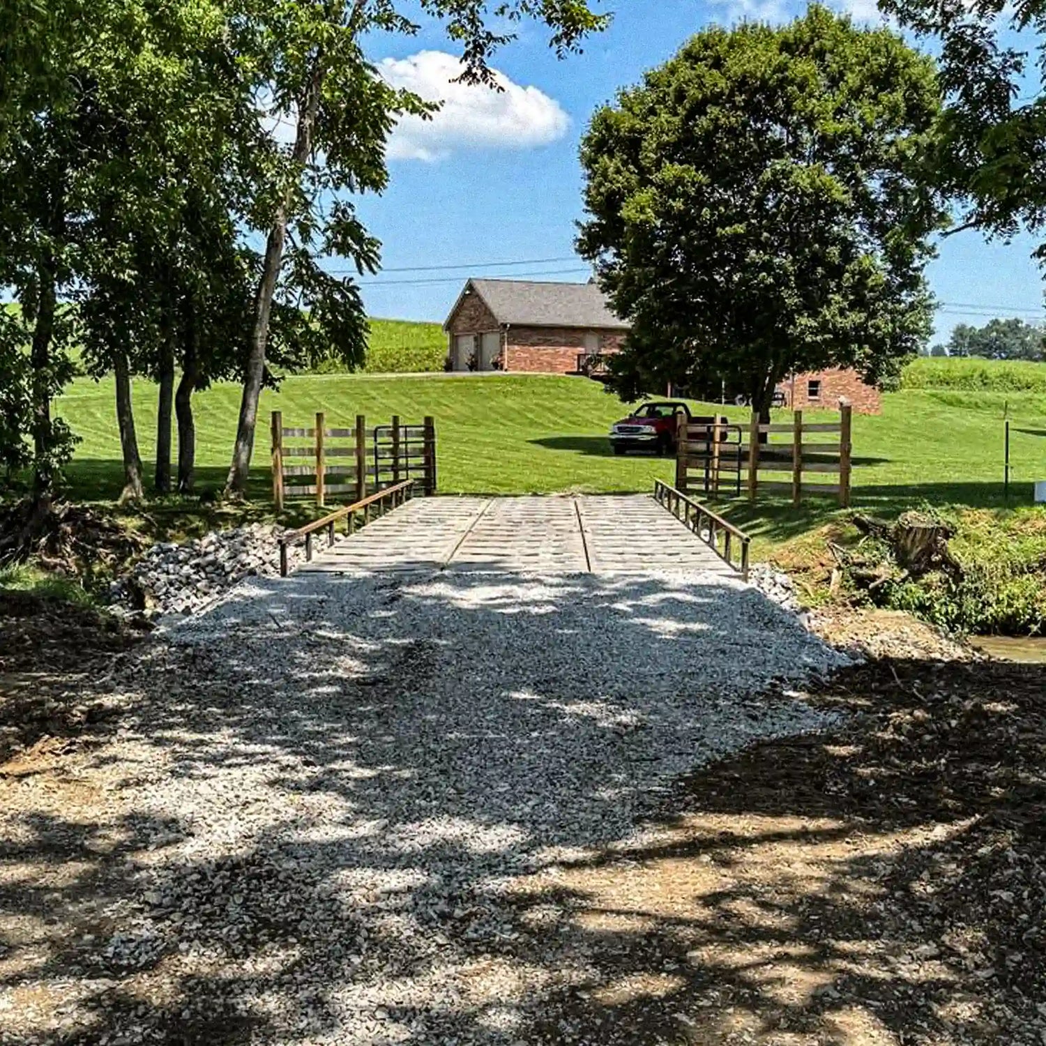 Front view of a modular steel residential bridge leading to a farmhouse property with gravel approaches and fenced entry.