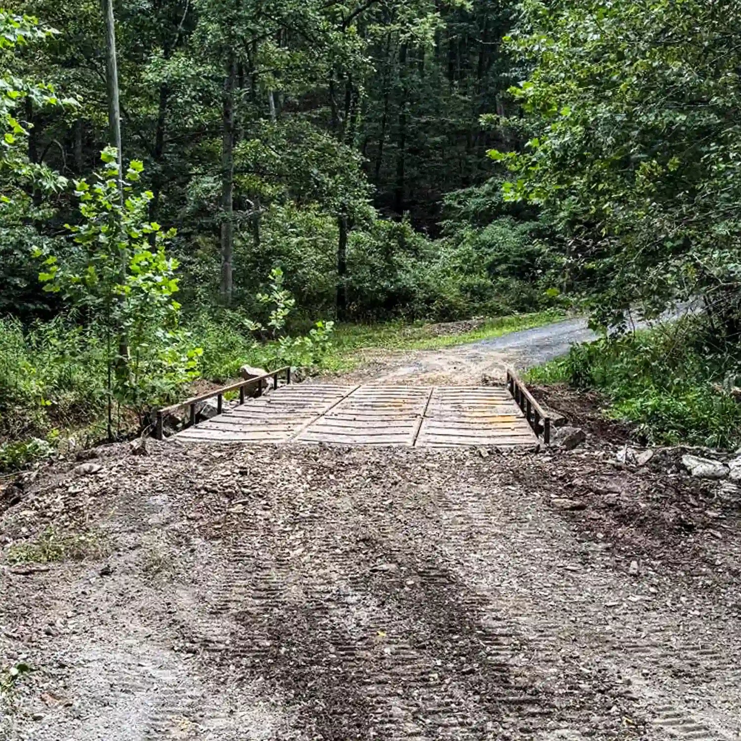 Front approach view of a residential steel bridge installed on a gravel driveway in a wooded setting.