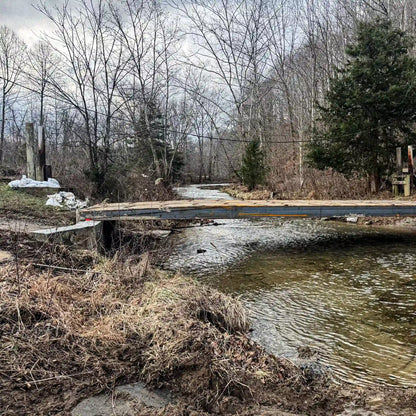 Side view of a modular steel residential bridge spanning a creek, connecting a rural driveway across a stream.