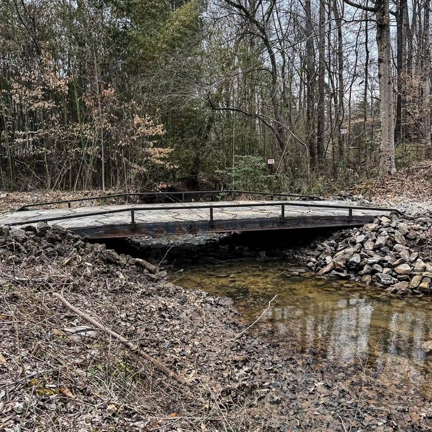Steel residential bridge installed over a shallow creek in a wooded area, with rock-lined banks and low side railings.
