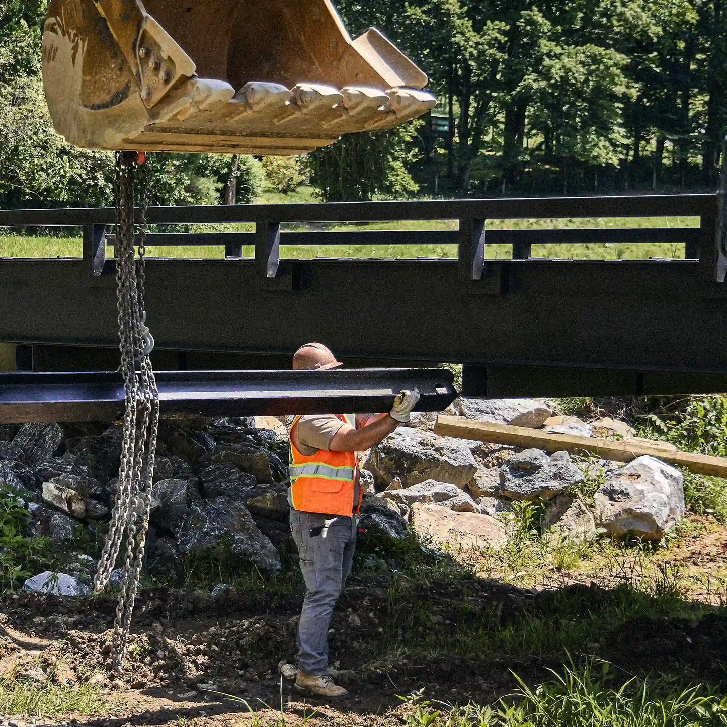 Excavator lifting a modular bridge section with chains over a creek crossing