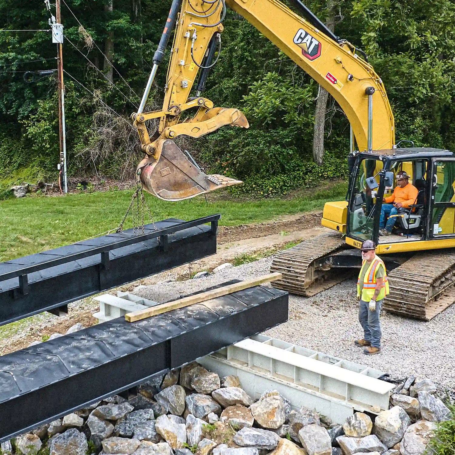 Excavator positioning a modular steel bridge section over a rock-lined creek crossing