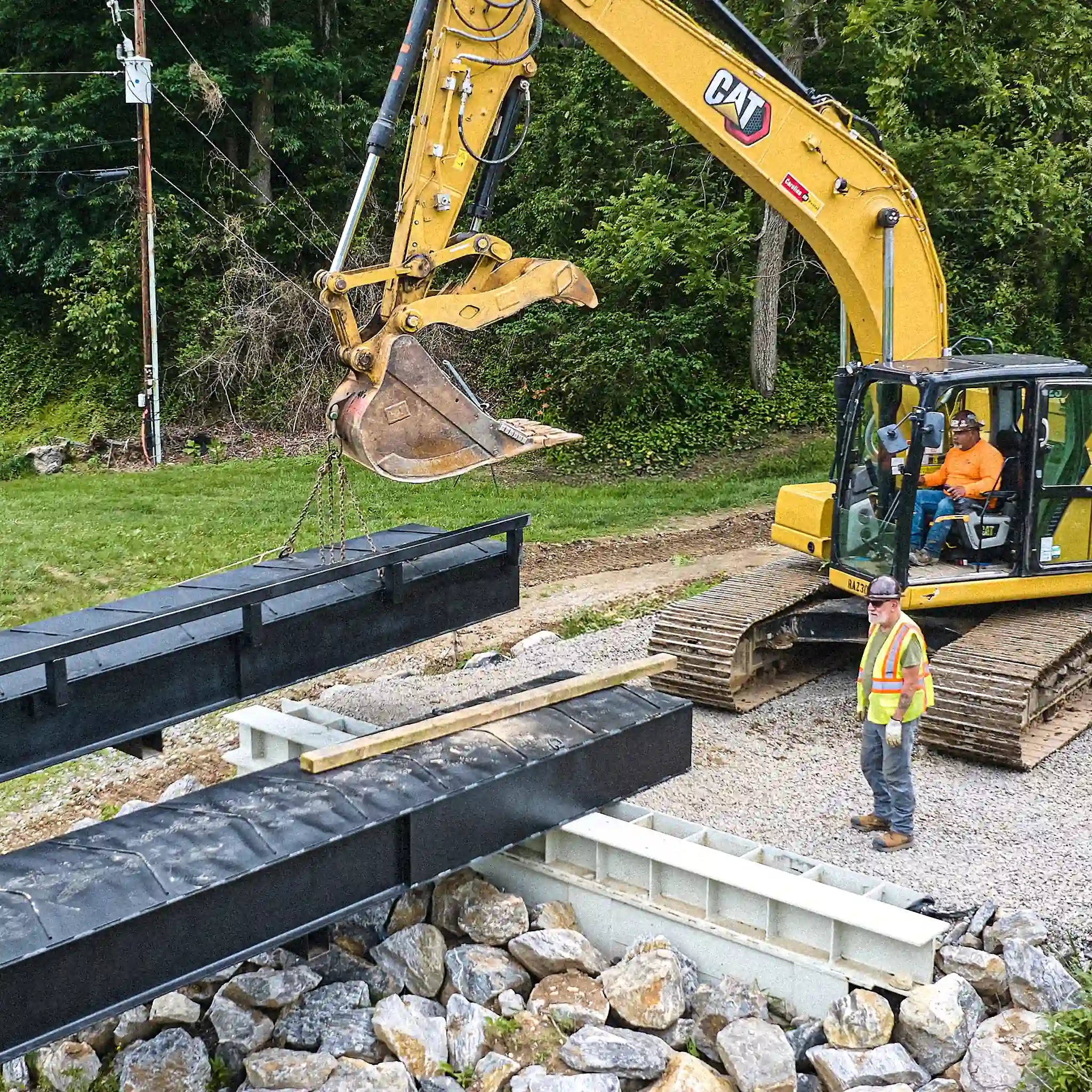 Excavator positioning a modular steel bridge section over a rock-lined creek crossing
