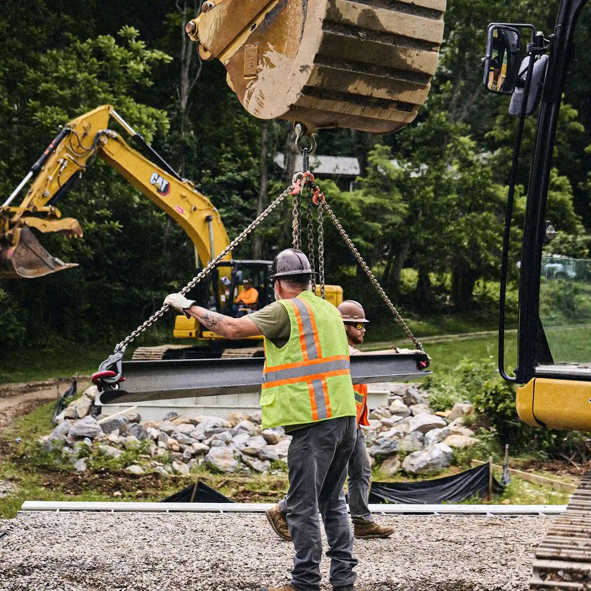 Worker guiding a modular bridge lift over a rock-lined creek crossing